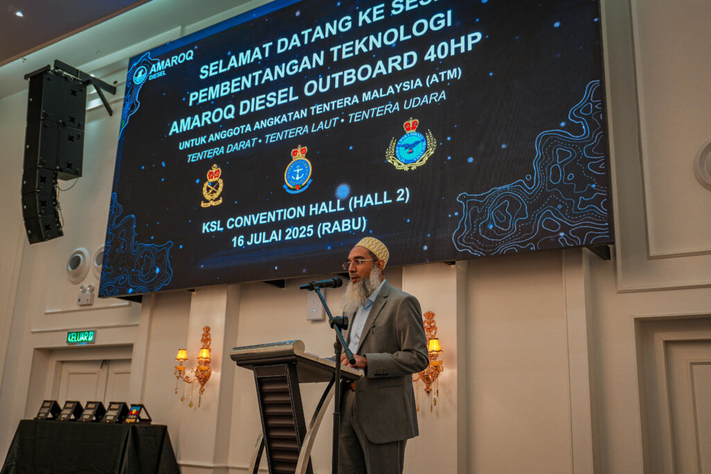 Speaker in a grey suit and traditional cap presenting at the podium during the AMAROQ Diesel Outboard 40HP Technology Presentation, held at KSL Convention Hall on 16 July 2025. A large LED screen behind displays event details for the Malaysian Armed Forces, featuring military insignias for the Army, Navy, and Air Force.